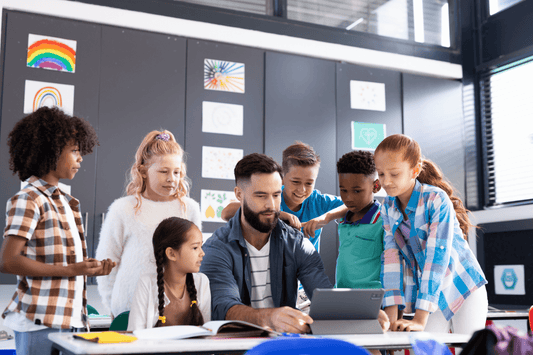 An educator seated at a classroom table holds a tablet while six students stand and sit around, looking at the screen together. Colorful artwork decorates the background walls.