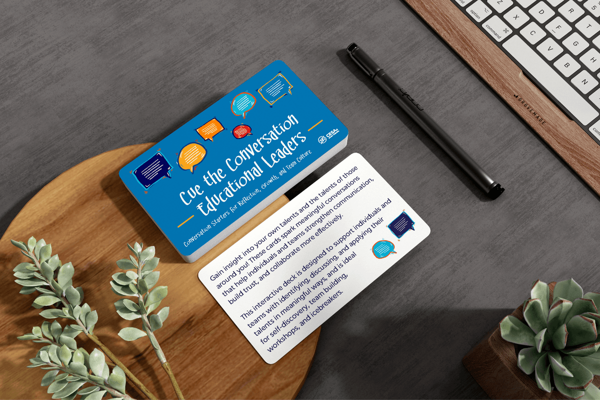 Blue “Conversation for Educational Leaders” cards sit on a wooden desk beside a pen, keyboard, and green plants, creating a thoughtful office setting.