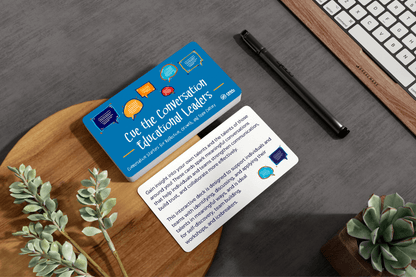 Blue “Conversation for Educational Leaders” cards sit on a wooden desk beside a pen, keyboard, and green plants, creating a thoughtful office setting.