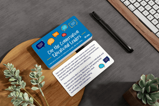 Blue “Conversation for Educational Leaders” cards sit on a wooden desk beside a pen, keyboard, and green plants, creating a thoughtful office setting.