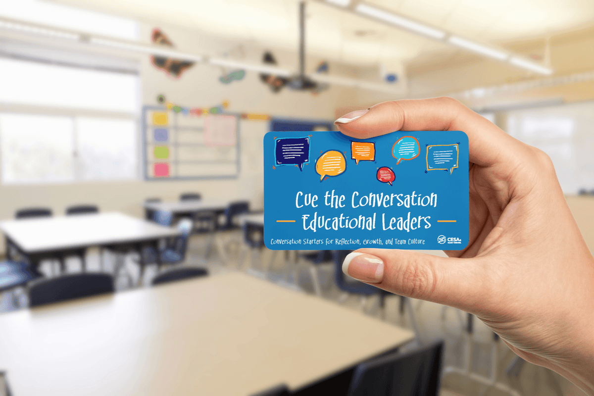Hand holds a blue card reading “Cue the Conversation: Educational Leaders” in a bright classroom with desks, chairs, and a welcoming tone.