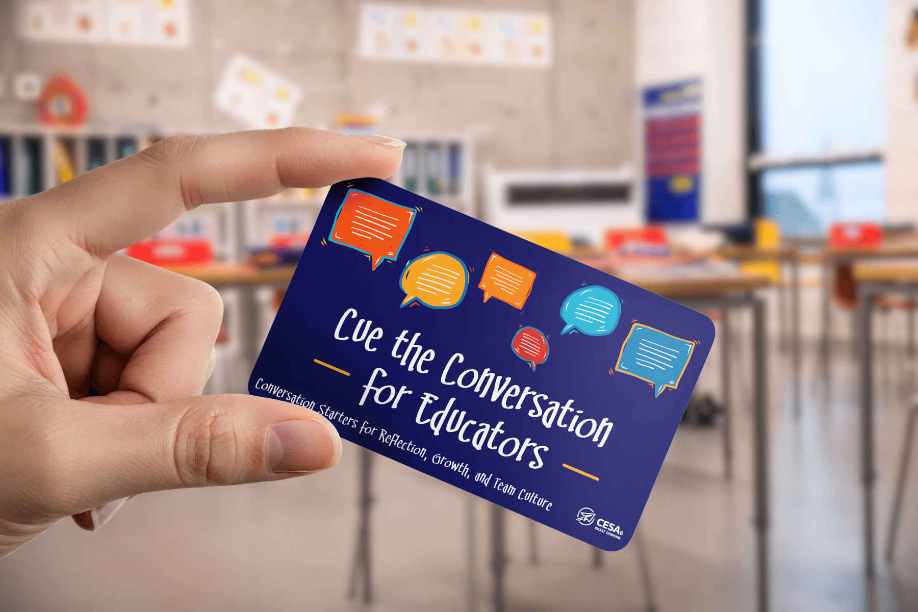 A hand holds a card with colorful speech bubbles and text: "Cue the Conversation for Educators." Blurred classroom background.