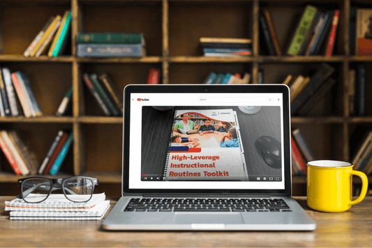 A laptop displays a video titled "High-Leverage Instructional Routines Toolkit" on a desk with glasses, notebooks, and a yellow mug. A bookshelf is in the background.