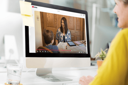 A person watches a YouTube video on a desktop screen, showing a smiling woman interacting with a child at a table. The setting is warm and educational.
