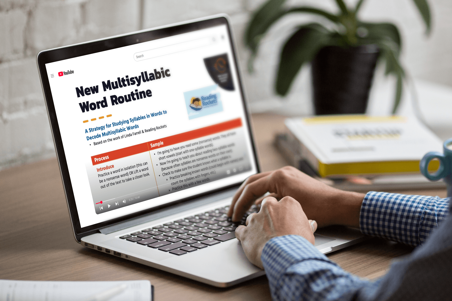 A person in a checkered shirt watches a YouTube video titled "New Multisyllabic Word Routine" on a laptop, with books and a plant nearby.