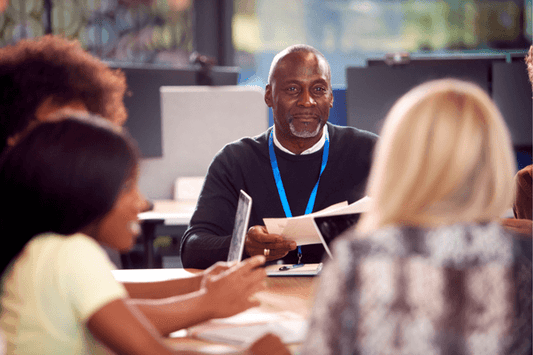 A group of adults gathers around a table in a modern office or classroom setting. At the center, a person wearing a dark sweater and a blue lanyard is holding a sheet of paper and appears to be leading a discussion. Others in the group are smiling, listening, or interacting with devices and documents. The atmosphere is collaborative and focused, with blurred computer monitors and windows in the background suggesting a professional or educational environment.