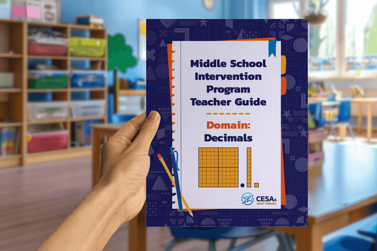 A hand holds a guide titled "Middle School Intervention Program Teacher Guide: Domain Decimals" with educational graphics. A classroom is blurred in the background.