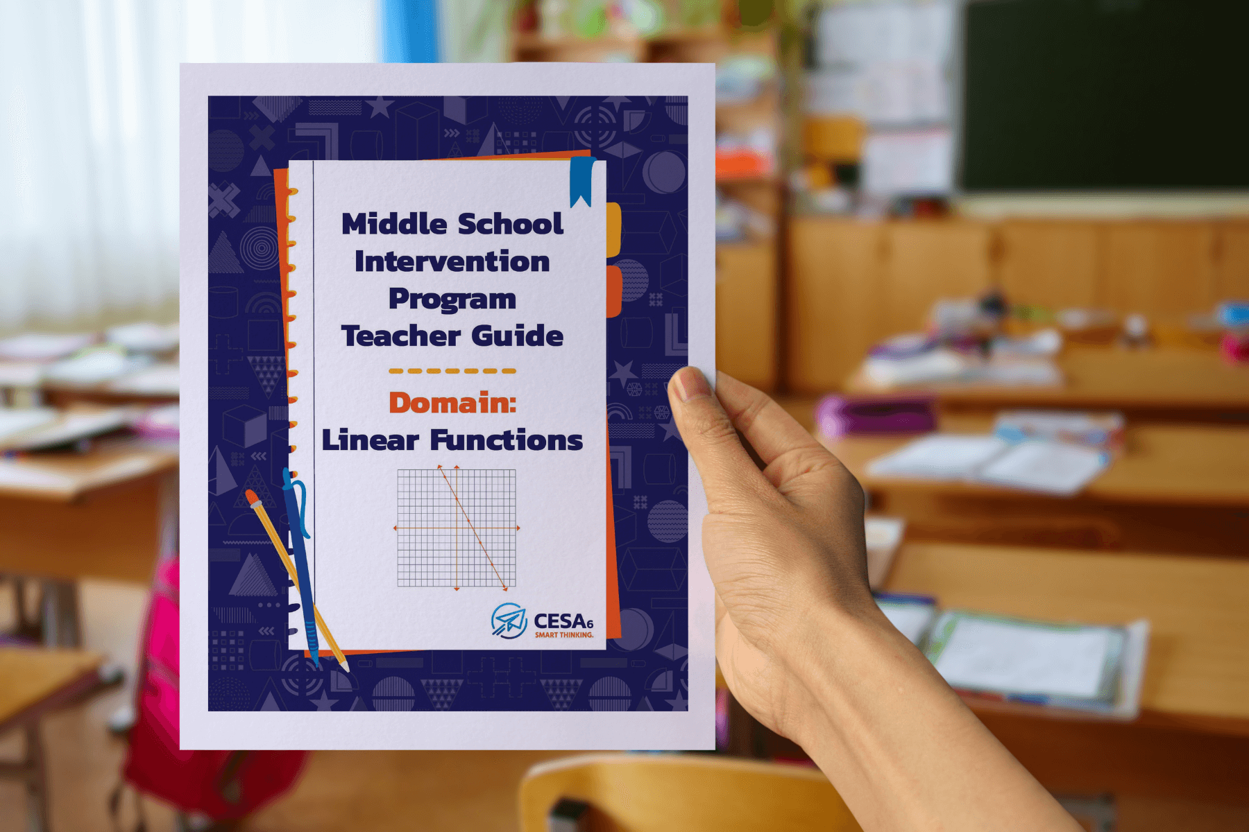 A person holds a booklet titled “Middle School Intervention Program Teacher Guide – Domain: Linear Functions”. The cover is decorated with classroom-themed graphics and features a coordinate grid with a downward-sloping line, representing a linear function. The background shows a classroom with wooden desks, papers, and school supplies.
