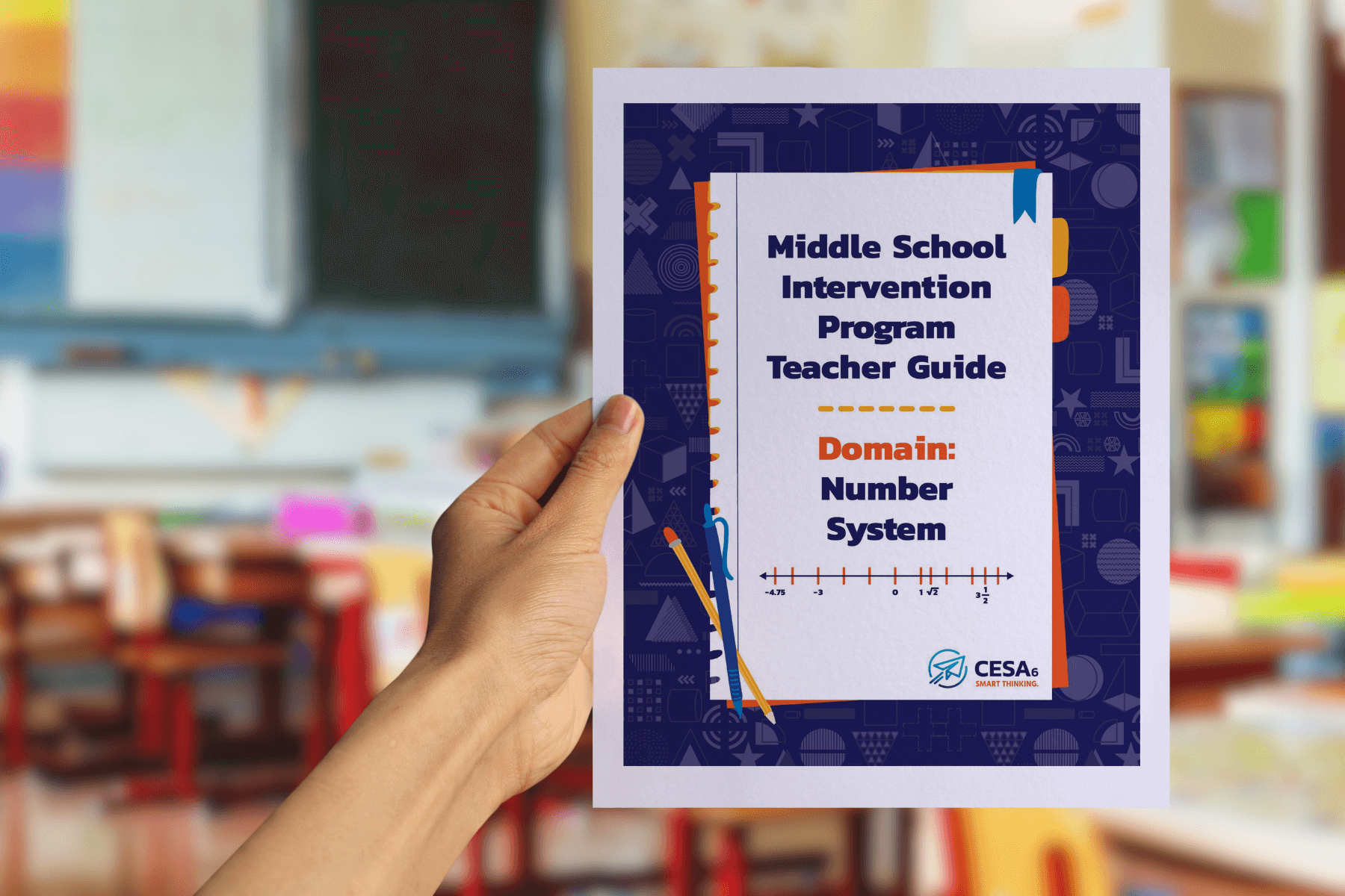 A person is holding up a booklet titled "Middle School Intervention Program Teacher Guide – Domain: Number System" in a classroom setting. The cover is designed with a dark blue patterned border, orange and blue accents, and features a number line graphic at the bottom. The blurred background shows colorful classroom decorations, bookshelves, and desks.