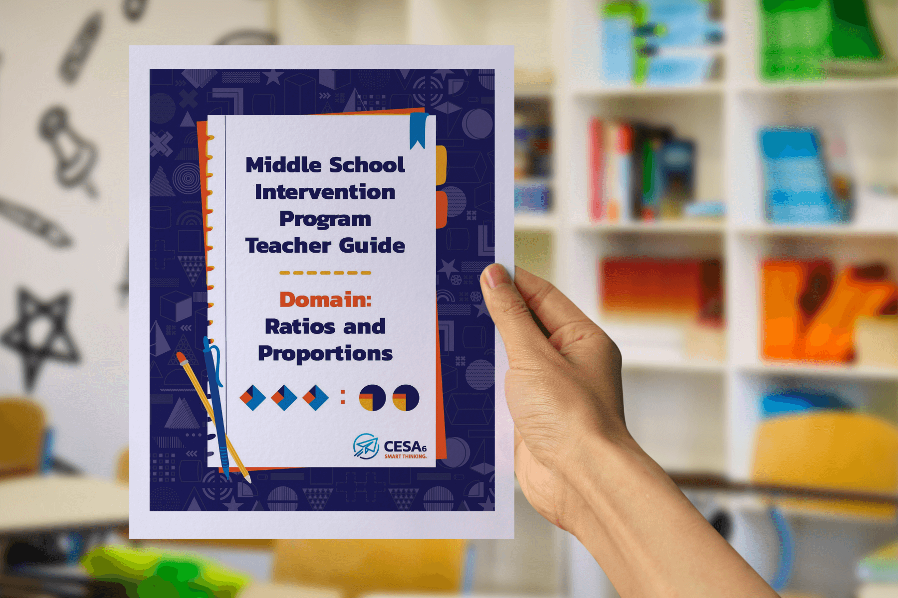A hand holds up a booklet titled Middle School Intervention Program Teacher Guide: Domain – Ratios and Proportions. The cover features colorful math-themed graphics, including geometric shapes and ratio symbols. In the background, blurred bookshelves and classroom chairs are visible, suggesting a school setting.
