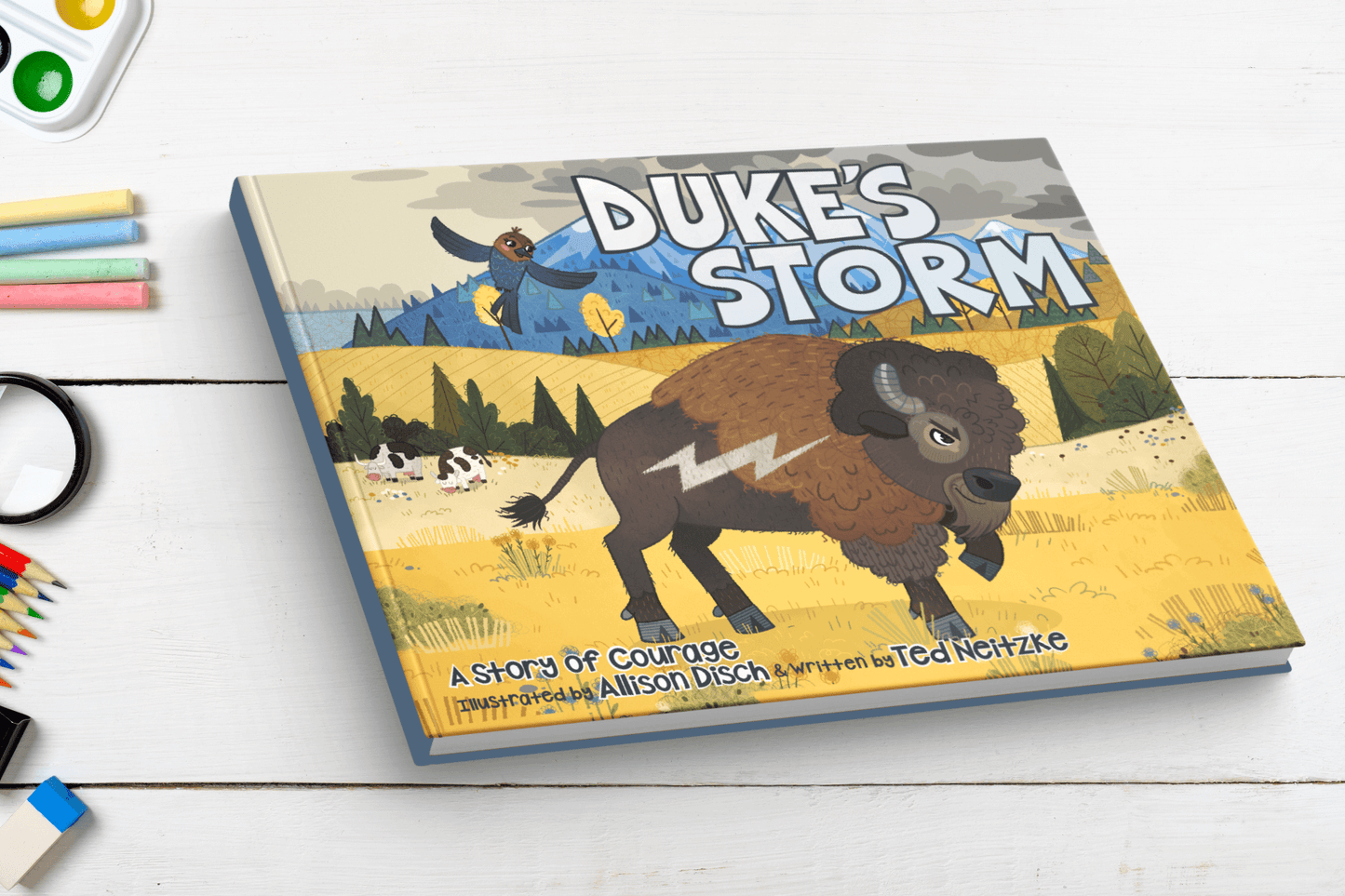 Hardcover book of 'Duke's Storm' placed on a white wooden table surrounded by art supplies, including colored pencils and watercolor paints. The cover features an illustrated bison, Duke with a lightning bolt symbol and a bird, Tootsie flying above, set in a scenic field with mountains in the background. The text reads 'A story of courage, illustrated by Allison Disch & written by Ted Neitzke.