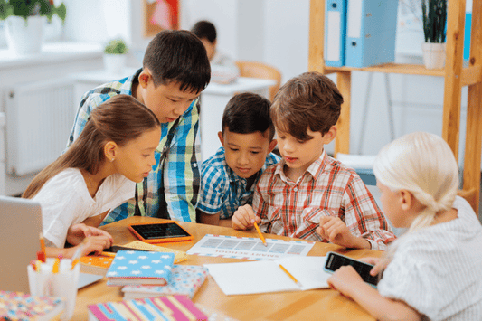 Five children are gathered around a table, working collaboratively on a project that includes papers, pencils, and tablets. They are leaning in closely and appear focused on the task, in a classroom setting.