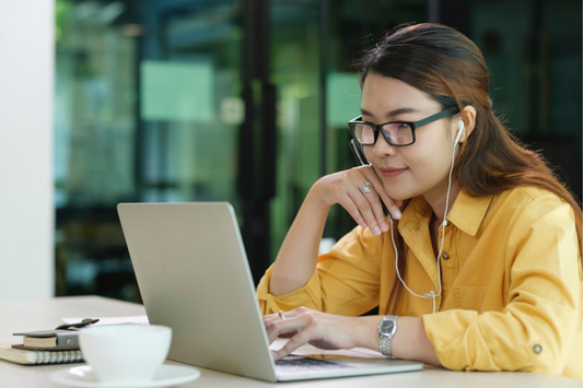 A woman in a yellow shirt sits at a table working on a laptop with earbuds in her ears. 