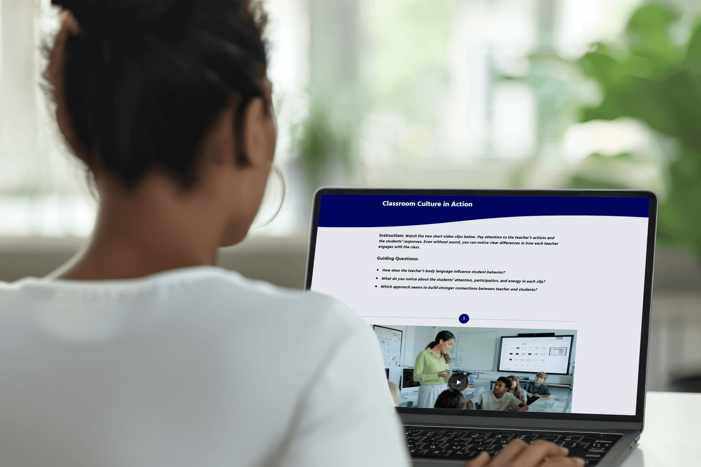A woman viewed from behind is using a laptop displaying an educational video titled "Classroom Culture in Action." She's in a cozy setting.