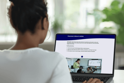 A woman viewed from behind is using a laptop displaying an educational video titled "Classroom Culture in Action." She's in a cozy setting.