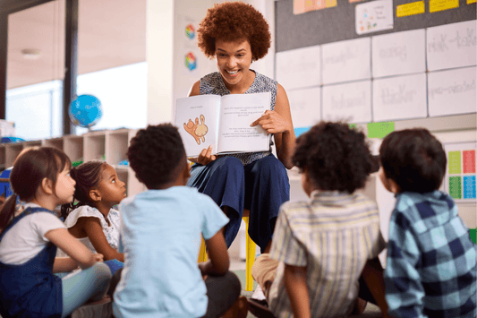 An educator seated on a small chair reads a picture book to a group of young students sitting on the floor in a classroom. The children are watching and listening attentively.