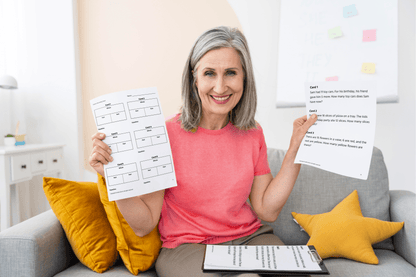 A smiling person with gray hair in a pink shirt holds up 1st Grade Lessons on a couch. Clipboard and yellow pillows are visible, conveying a positive mood.