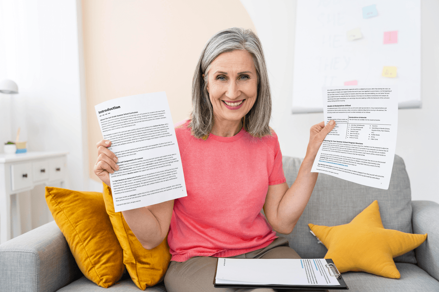 A smiling person with gray hair in a pink shirt holds up 2nd Grade Lessons on a couch. Clipboard and yellow pillows are visible, conveying a positive mood.