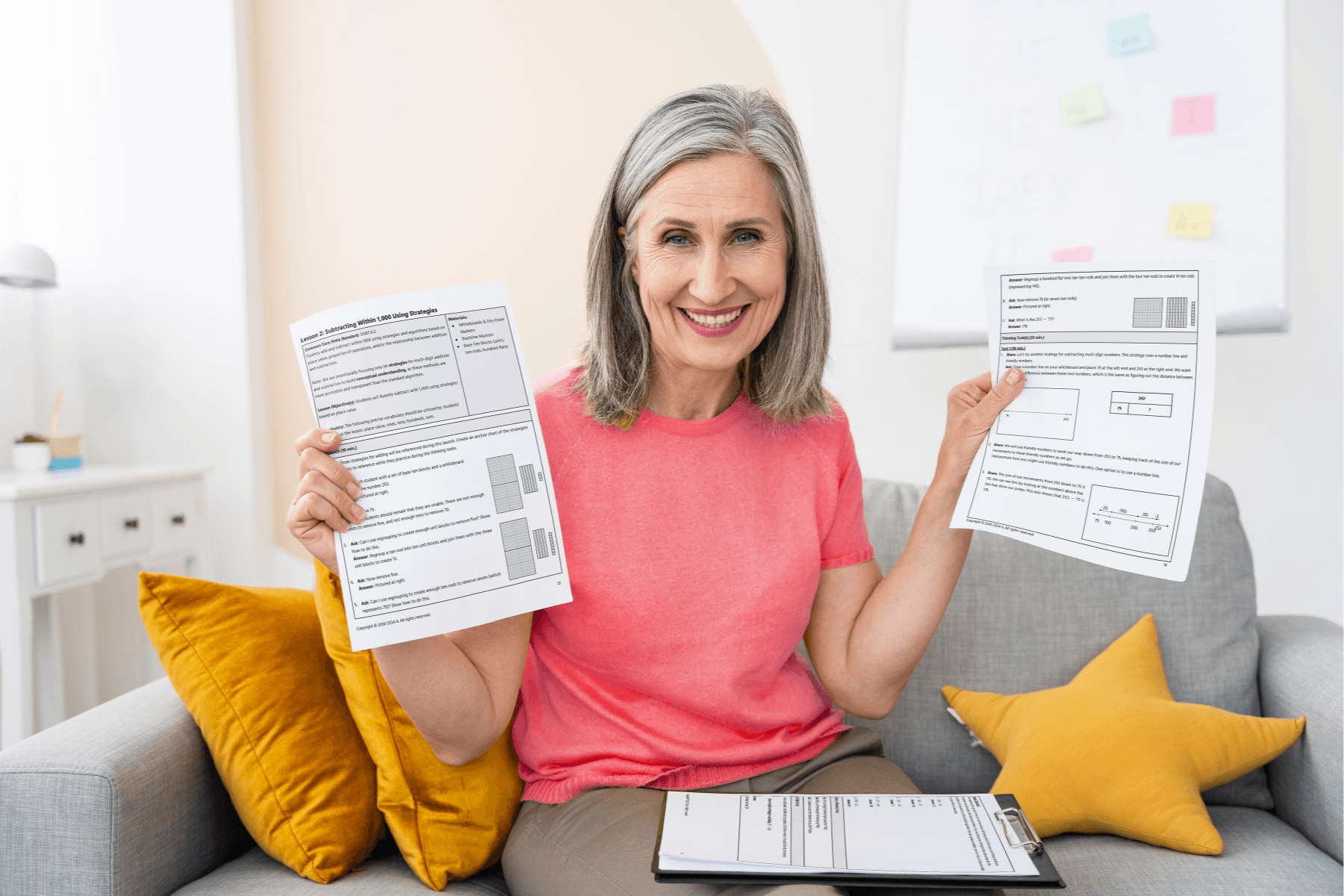 A smiling person with gray hair in a pink shirt holds up 3rd Grade Lessons on a couch. Clipboard and yellow pillows are visible, conveying a positive mood.