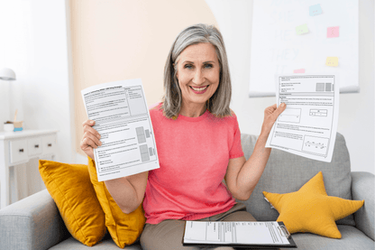 A smiling person with gray hair in a pink shirt holds up 3rd Grade Lessons on a couch. Clipboard and yellow pillows are visible, conveying a positive mood.