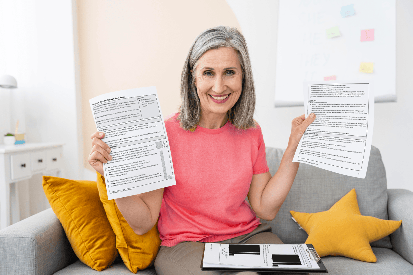 A smiling person with gray hair in a pink shirt holds up 4th Grade Lessons on a couch. Clipboard and yellow pillows are visible, conveying a positive mood.