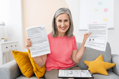 A smiling person with gray hair in a pink shirt holds up 5th grade Lessons on a couch. Clipboard and yellow pillows are visible, conveying a positive mood.