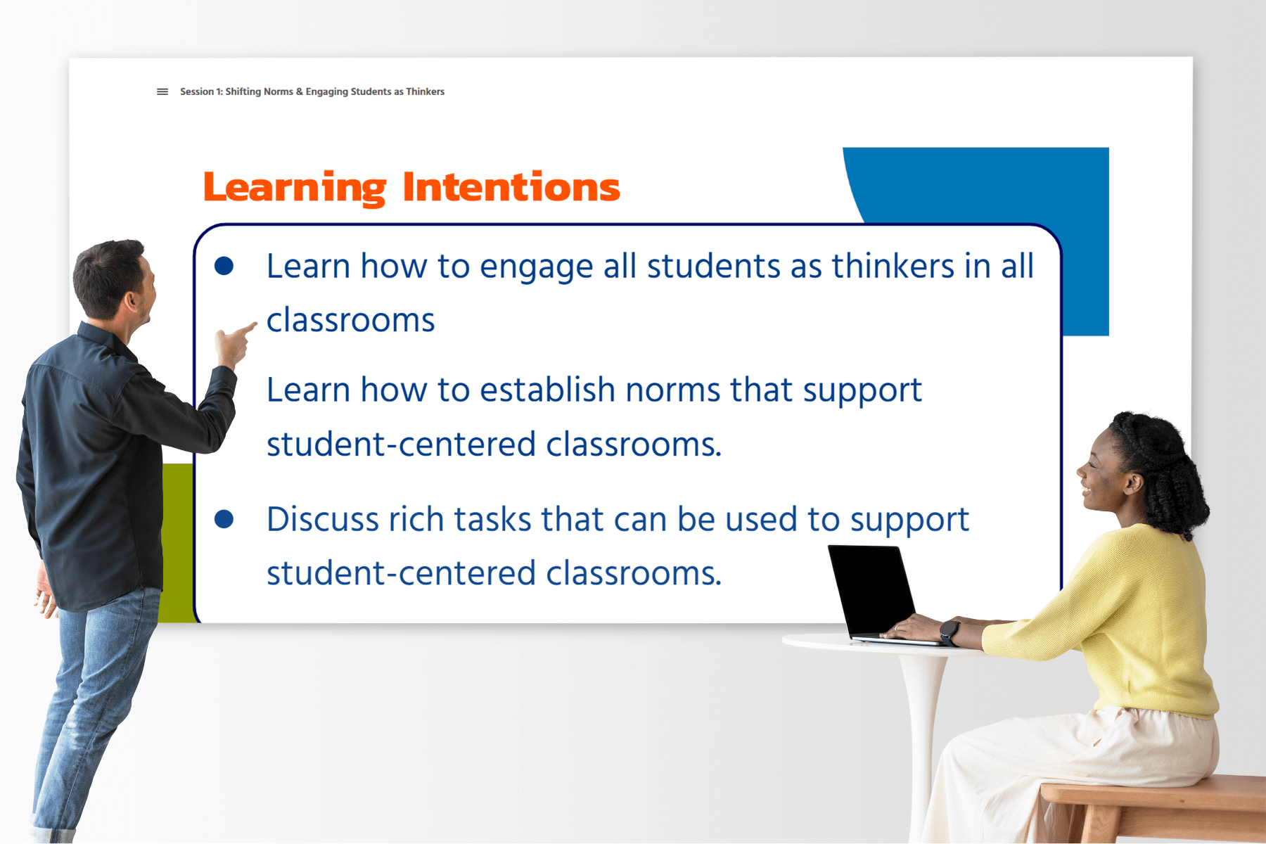 Two educators are interacting with a large projected slide titled “Learning Intentions.” The slide outlines three goals: engaging all students as thinkers, establishing student-centered norms, and discussing rich tasks that promote student-centered classrooms. The educator on the left is pointing toward the slide, while the educator on the right, seated at a table with a laptop, smiles and looks engaged.