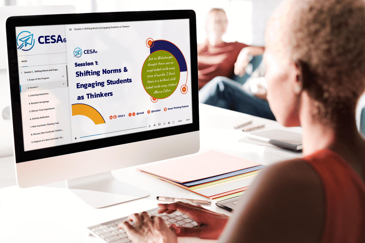 An educator is seated at a desk engaging with a professional learning module on a desktop computer. The screen displays a session titled “Shifting Norms & Engaging Students as Thinkers” from CESA 6, including a quote by Marva Collins. The desktop interface shows a structured learning menu with modules, and colorful folders and pens are organized nearby.