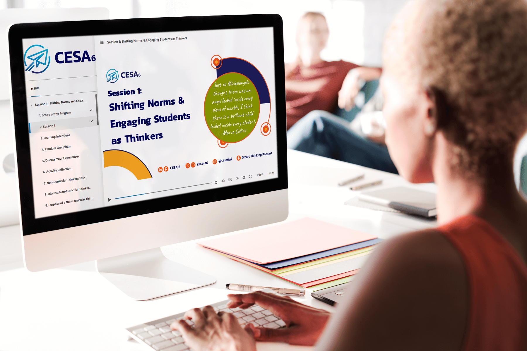An educator is seated at a desk engaging with a professional learning module on a desktop computer. The screen displays a session titled “Shifting Norms & Engaging Students as Thinkers” from CESA 6, including a quote by Marva Collins. The desktop interface shows a structured learning menu with modules, and colorful folders and pens are organized nearby.