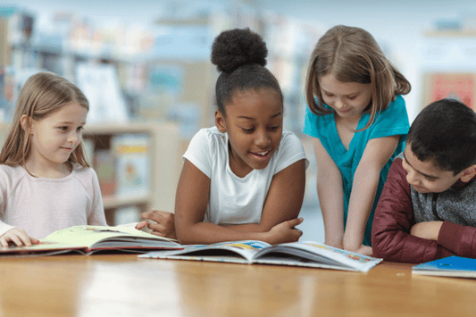 Four young students sit and stand around a table in a library, smiling and reading picture books together.