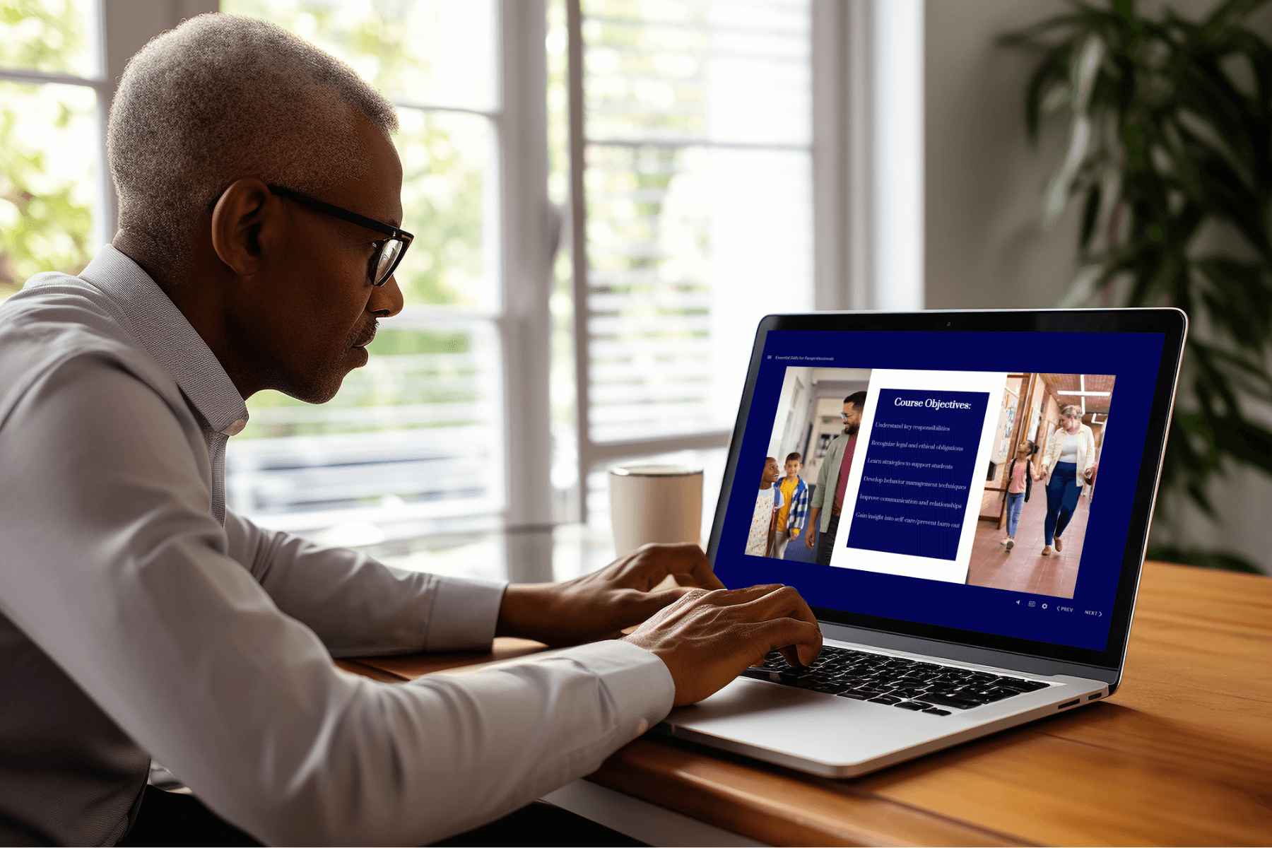 A person with short gray hair and glasses is sitting at a wooden desk, working on a laptop. The laptop screen displays an online course titled "Course Objectives," with two images of adults guiding children in a school hallway on either side of the text. The individual is attentively typing or navigating the course. A white coffee cup is on the desk, and large windows with sunlight and greenery outside provide a bright, natural background.