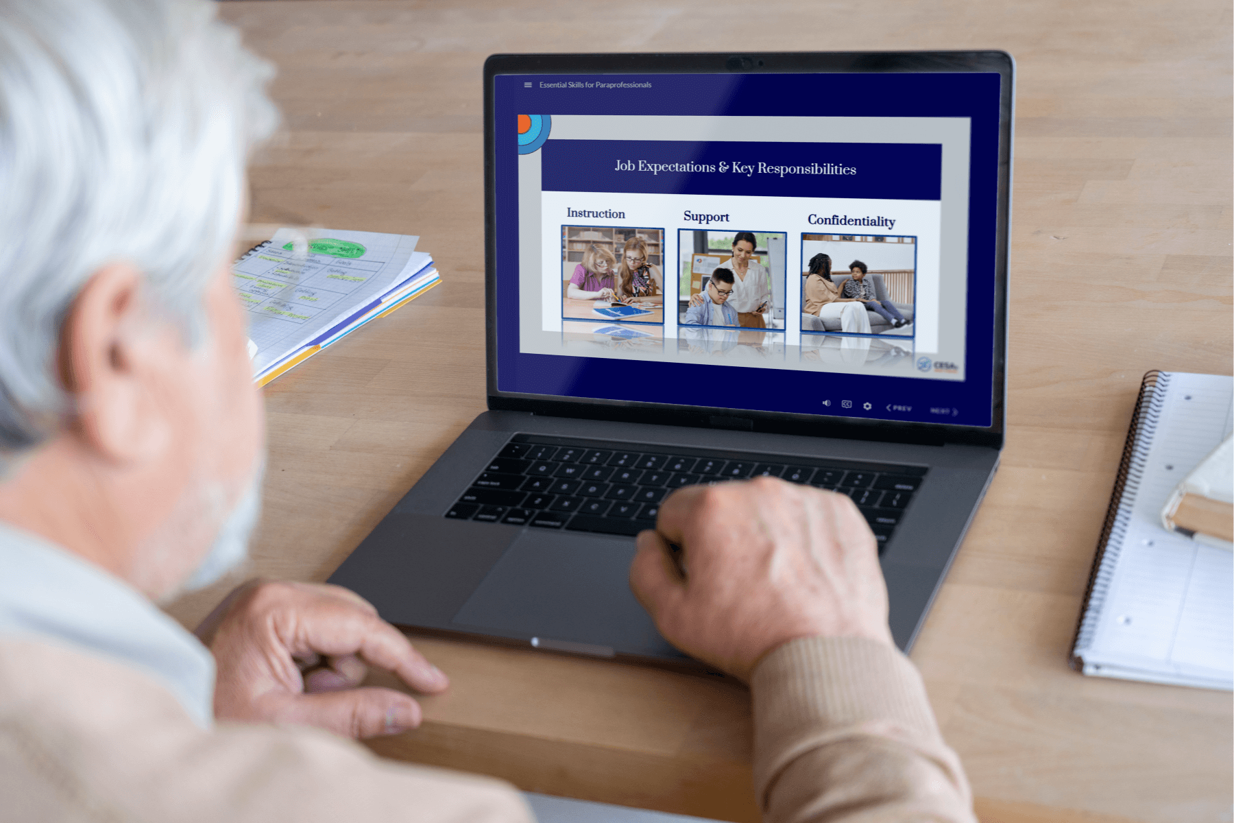 An older adult sits at a table using a laptop. On the laptop screen is a course slide titled "Job Expectations & Key Responsibilities" from the Essential Skills for Paraprofessionals training. The slide shows three sections labeled "Instruction," "Support," and "Confidentiality," each accompanied by a photo of an educator working with students.