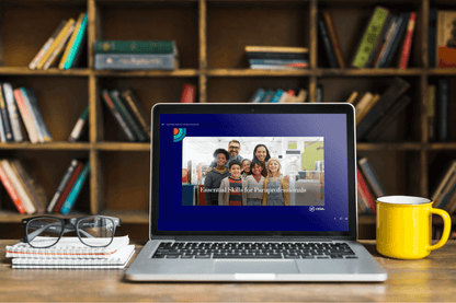 A laptop sits on a wooden desk in front of a bookshelf filled with books. On the laptop screen is the opening slide of the Essential Skills for Paraprofessionals course, displaying a smiling group of educators and students.