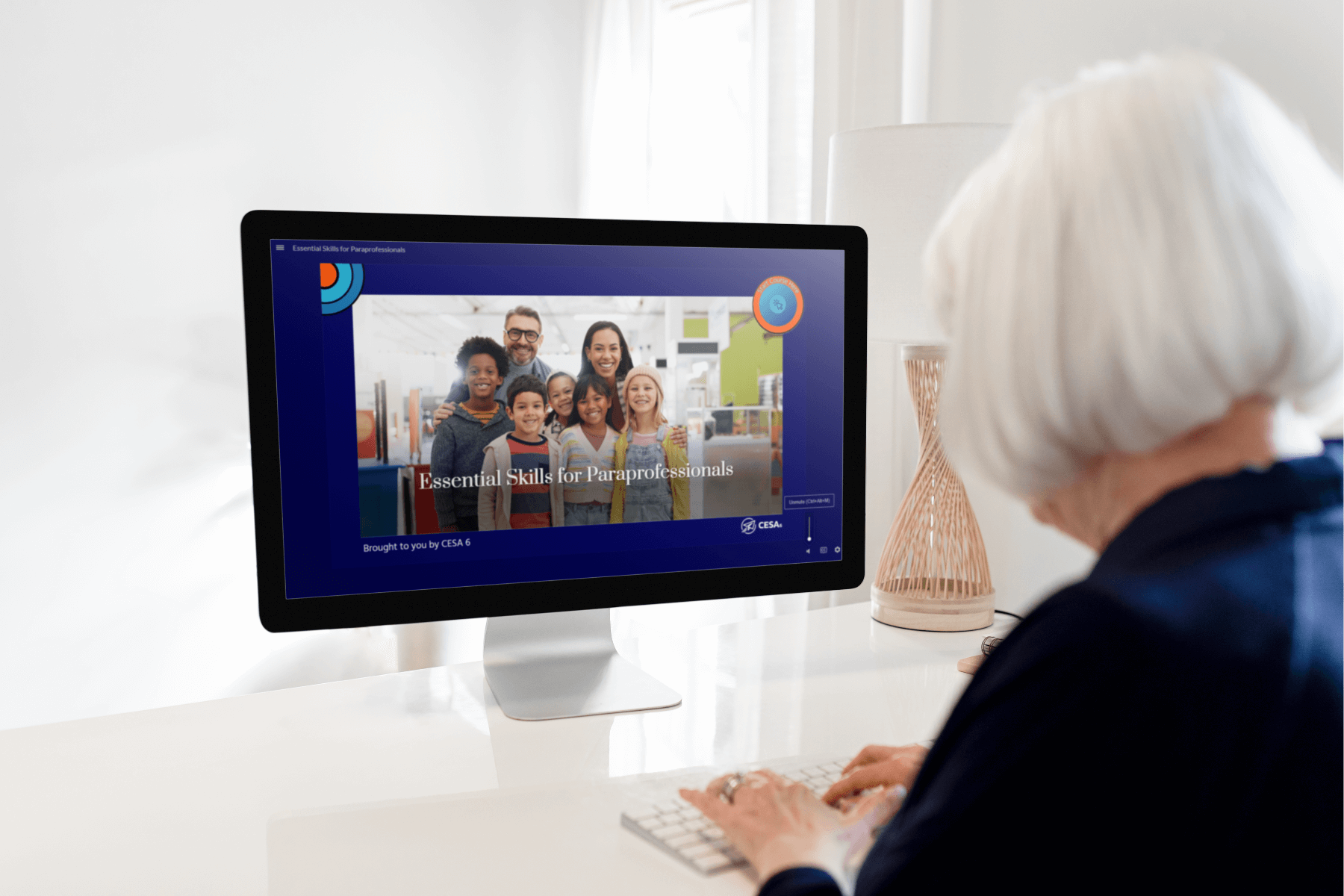 An older adult with short white hair sits at a desk using a desktop computer. On the screen is the opening slide of the Essential Skills for Paraprofessionals course, showing a group of smiling educators and students. The background is bright with modern classroom decor, and the course is branded by CESA 6.