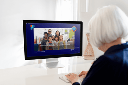 An older adult with short white hair sits at a desk using a desktop computer. On the screen is the opening slide of the Essential Skills for Paraprofessionals course, showing a group of smiling educators and students. The background is bright with modern classroom decor, and the course is branded by CESA 6.