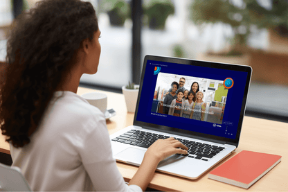 A person with long curly hair, wearing a white shirt, sits at a desk using a laptop. The laptop screen displays an online course titled "Essential Skills for Paraprofessionals," featuring a group photo of smiling adults and children. The course is labeled as being provided by CESA 6. 