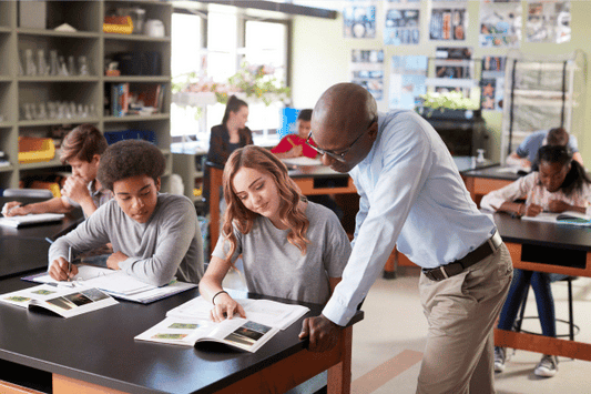 A high school science classroom where students are seated at lab tables, engaged in reading textbooks and taking notes. In the foreground, a teacher is leaning over to assist a student who is pointing to something in her book. 