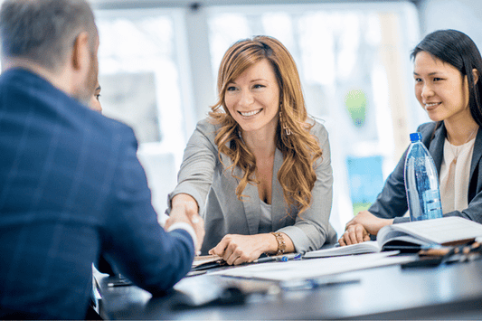 A group of professionals sits at a table in a well-lit office or meeting room. One individual extends a handshake across the table while smiling, suggesting the start or conclusion of a meeting. Open notebooks, a laptop, and a water bottle are visible on the table, indicating a collaborative or business setting.