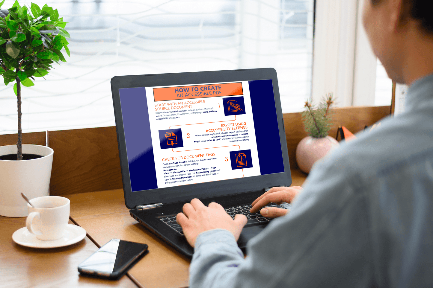 A person sits at a wooden desk using a laptop. The screen displays a guide titled "How to Create an Accessible PDF," with colorful icons and text. Nearby are a potted plant and a white mug, creating a cozy work environment.