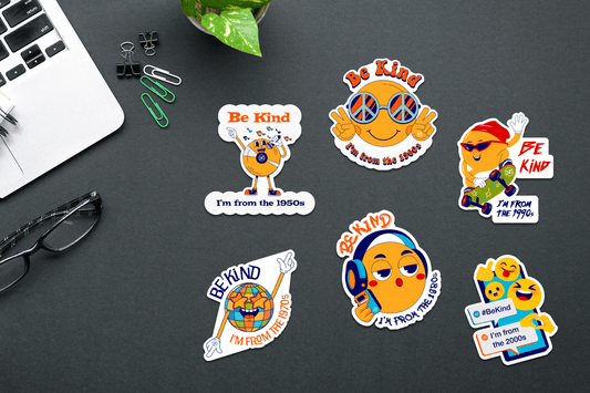 A collection of colorful vinyl stickers on a dark desk, each featuring a cartoon emoji-style character representing a different decade from the 1950s to the 2000s. Each sticker includes the phrase “Be Kind” with era-specific visual elements like sunglasses, a globe, a cassette player, or a smartphone. Office supplies, a laptop, a plant, and glasses are visible around the stickers.