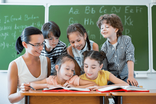 Teacher sitting at a desk with a group of young students, all looking at an open book together in a classroom with chalkboards in the background.