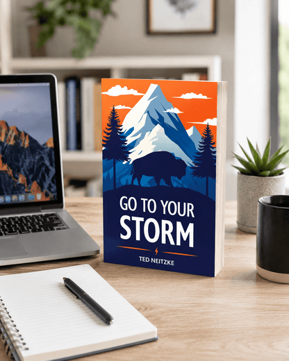 A book titled "Go to Your Storm" stands on a wooden desk with a laptop, notebook, pen, and mug nearby. The cover features a mountain, trees, and a bison, evoking a sense of adventure and resilience.