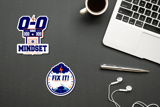 Two motivational leadership stickers displayed on a dark desk beside a laptop, coffee cup, pen, and earbuds. One sticker features a 0–0 scoreboard with the words “0-0 Mindset,” while the other features a storm cloud, lightning bolt, mountains, and buffalo with the words “Fix It!”