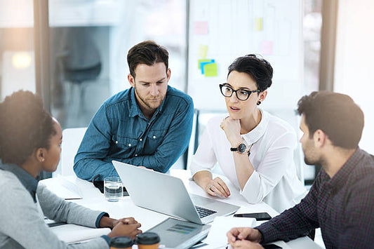 The image shows a group of four professionals having a collaborative discussion around a table. They are engaged with a laptop, and one person is speaking while the others listen attentively. 