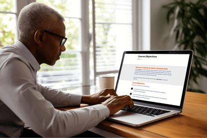 A person wearing glasses is focused on a laptop screen displaying course objectives while sitting at a wooden desk with a white mug, in a bright room.