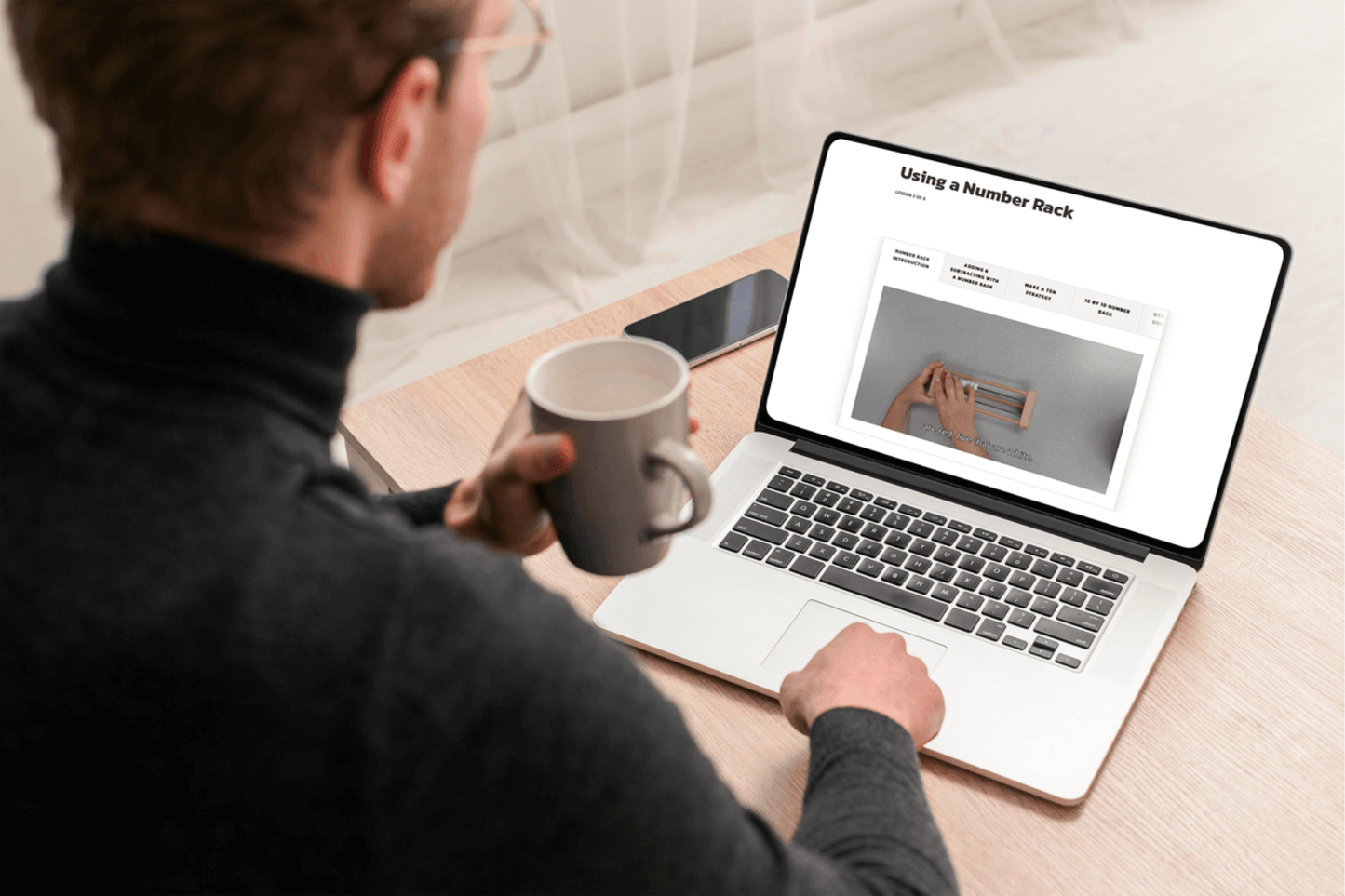 Man in glasses, holding coffee mug, looks at laptop screen displaying a webpage titled "Using a Number Rack." Desk, phone, and neutral tone setting.