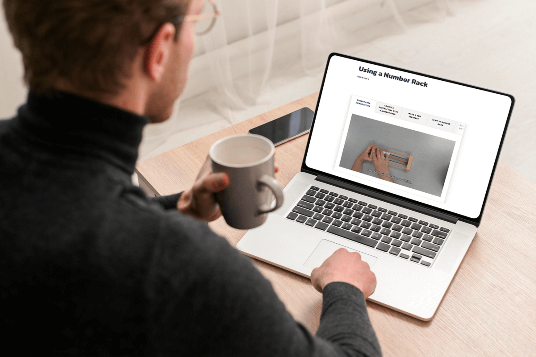 Man in glasses, holding coffee mug, looks at laptop screen displaying a webpage titled "Using a Number Rack." Desk, phone, and neutral tone setting.