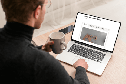 Man in glasses, holding coffee mug, looks at laptop screen displaying a webpage titled "Using a Number Rack." Desk, phone, and neutral tone setting.