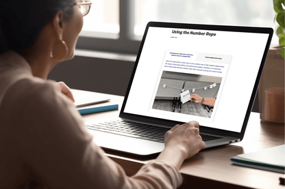 A person with glasses is sitting at a desk, looking at a laptop screen displaying an lesson titled "Using the Number Rope," focused on education.