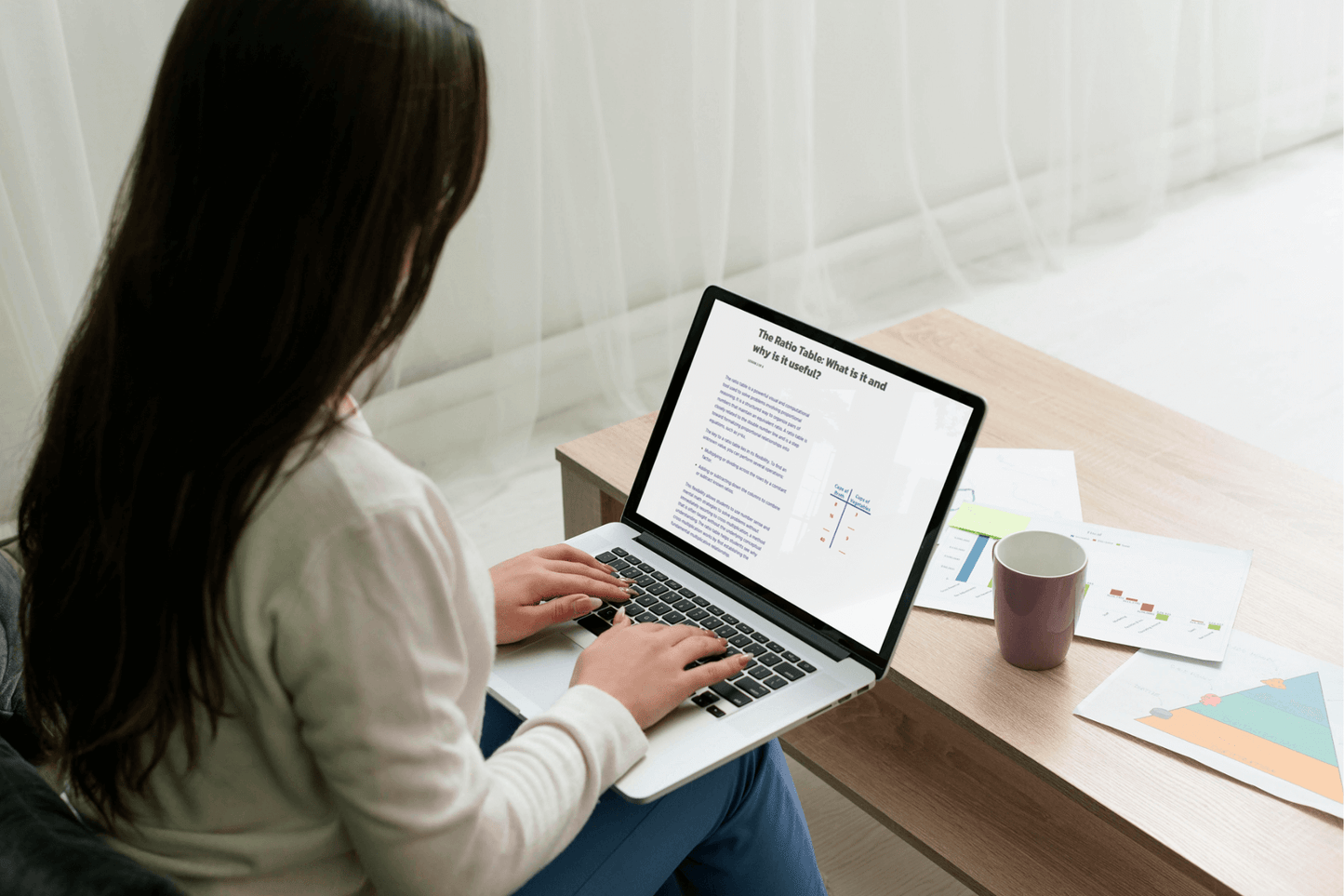 A woman sits on a couch, typing on a laptop displaying text and graphs. The scene includes a coffee mug and scattered charts on a wooden table. Cozy and focused atmosphere.